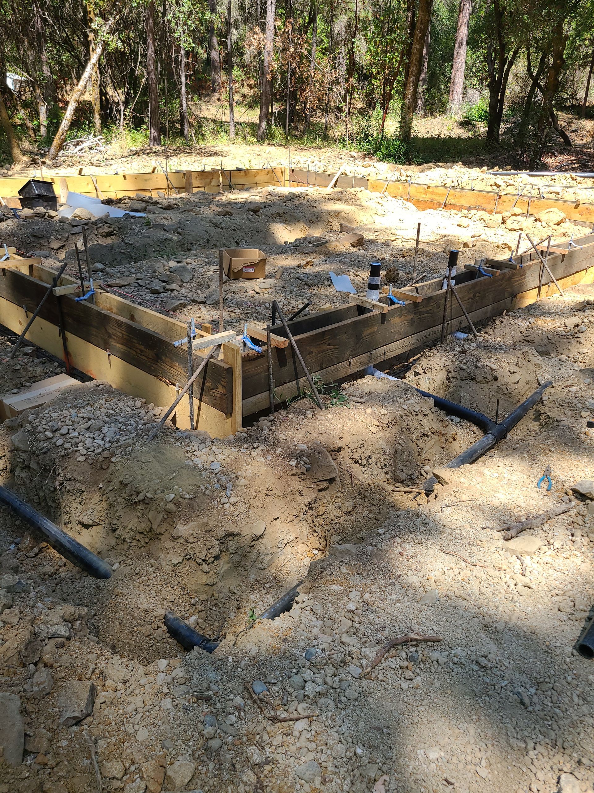Wooden frame for a foundation, with gravel and metal bars, in a dirt setting with trees.