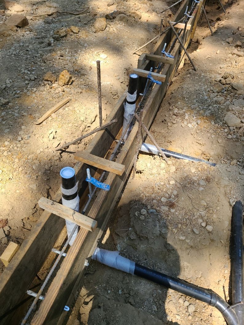 Plumbing pipes installed in a wooden trench, held in place with wooden supports, set in dirt.