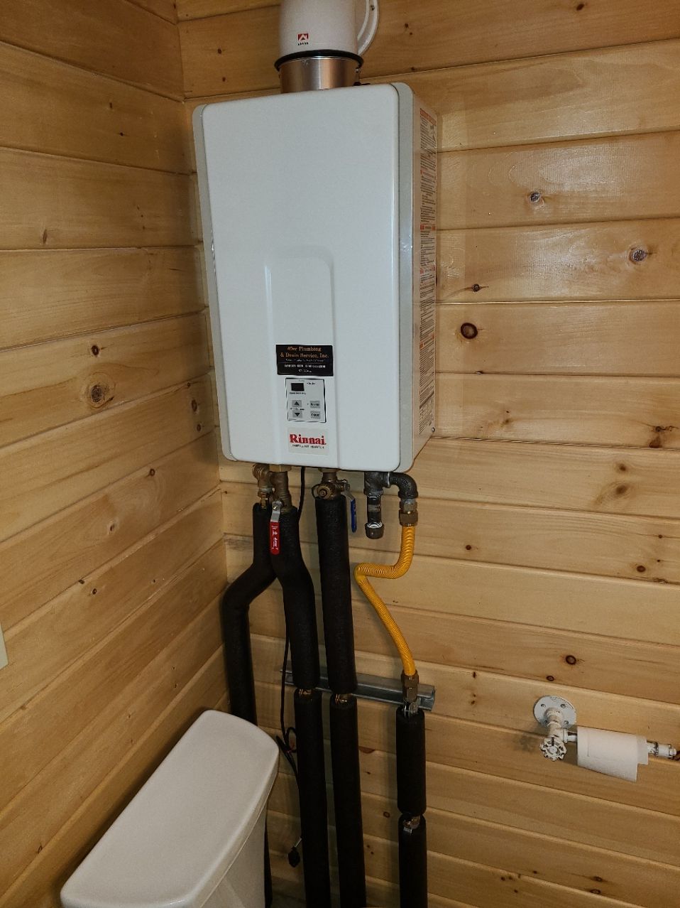 White wall-mounted water heater on wooden paneling, with black pipes and yellow gas line.