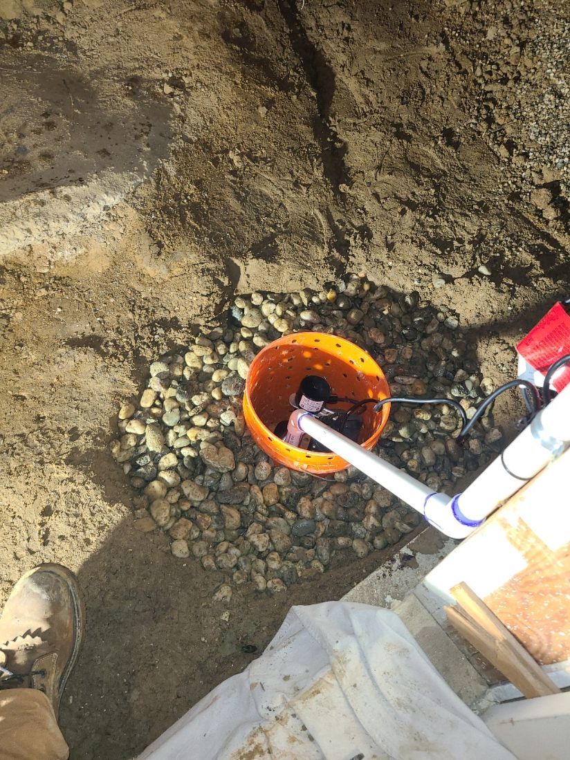 Orange bucket with sump pump in gravel, next to a white pipe. A person's boot is in the lower left corner.
