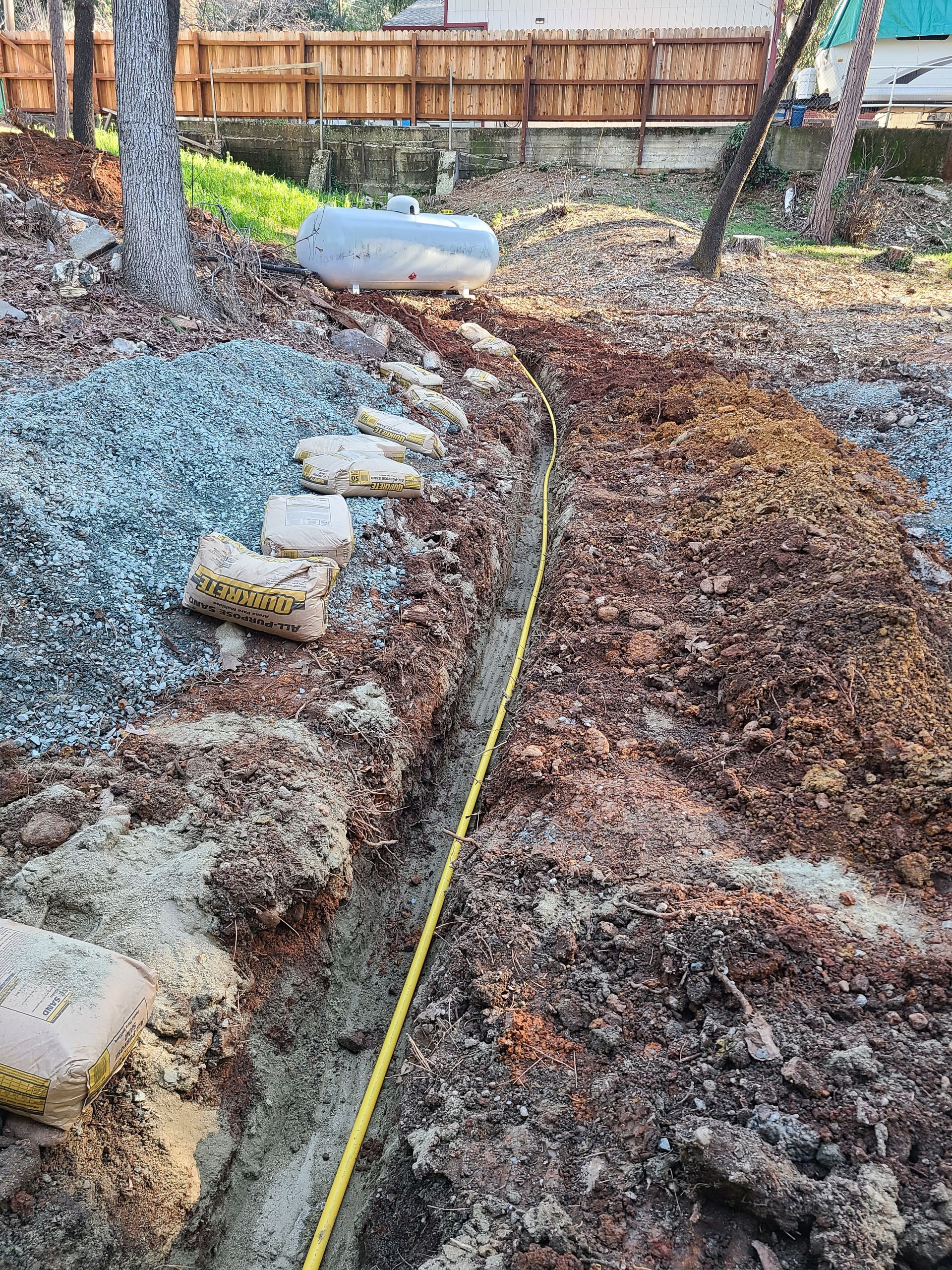 A trench dug in a yard, marked with a yellow tape, near stepping stones, and a propane tank.