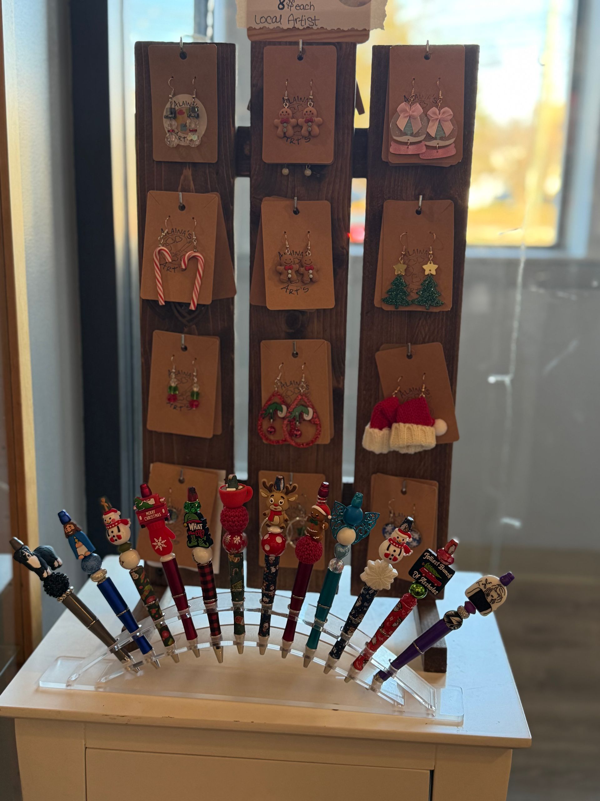 Display of handmade Christmas earrings and decorative pens on a white shelf.