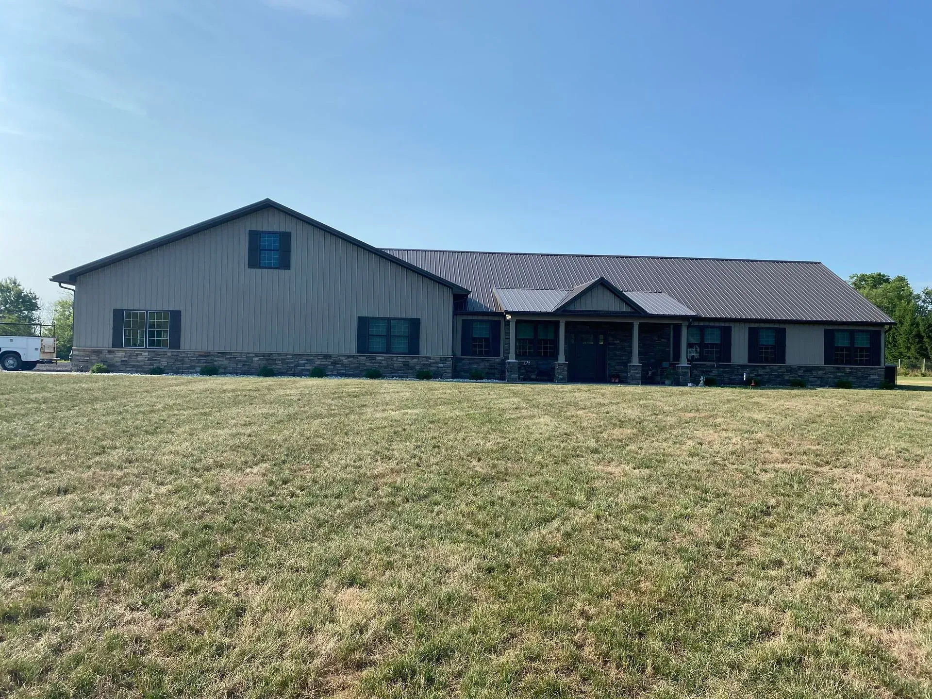 Tan house with a metal roof, stone foundation, and a porch, set in a grassy yard under a blue sky.