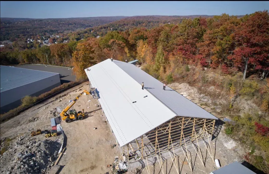 Construction of a large metal-roofed building on a hillside; autumn foliage in the background.