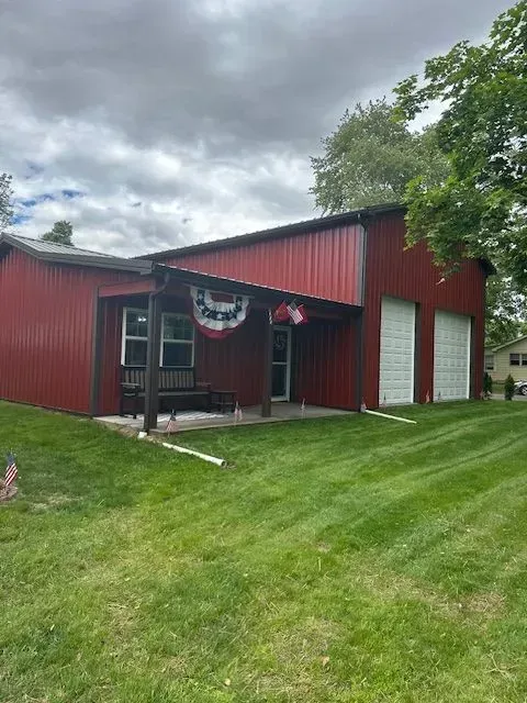 Red metal building with garage doors, porch, and American flag bunting on a lawn under a cloudy sky.