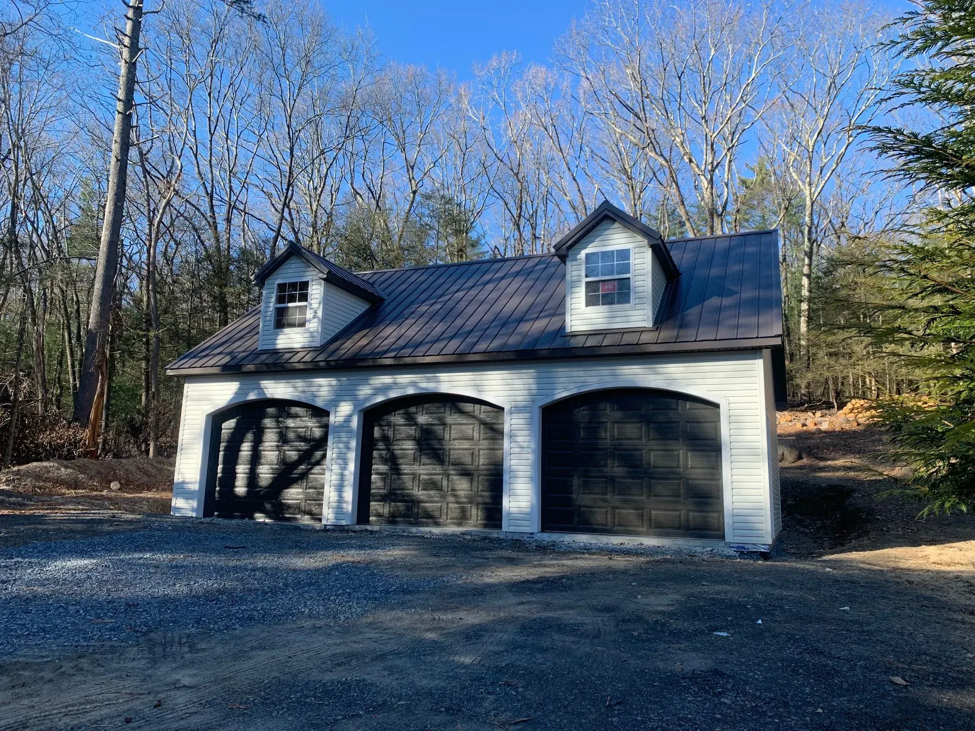 Three-car garage with dark doors, two dormers, and a brown metal roof. Set in a wooded area.