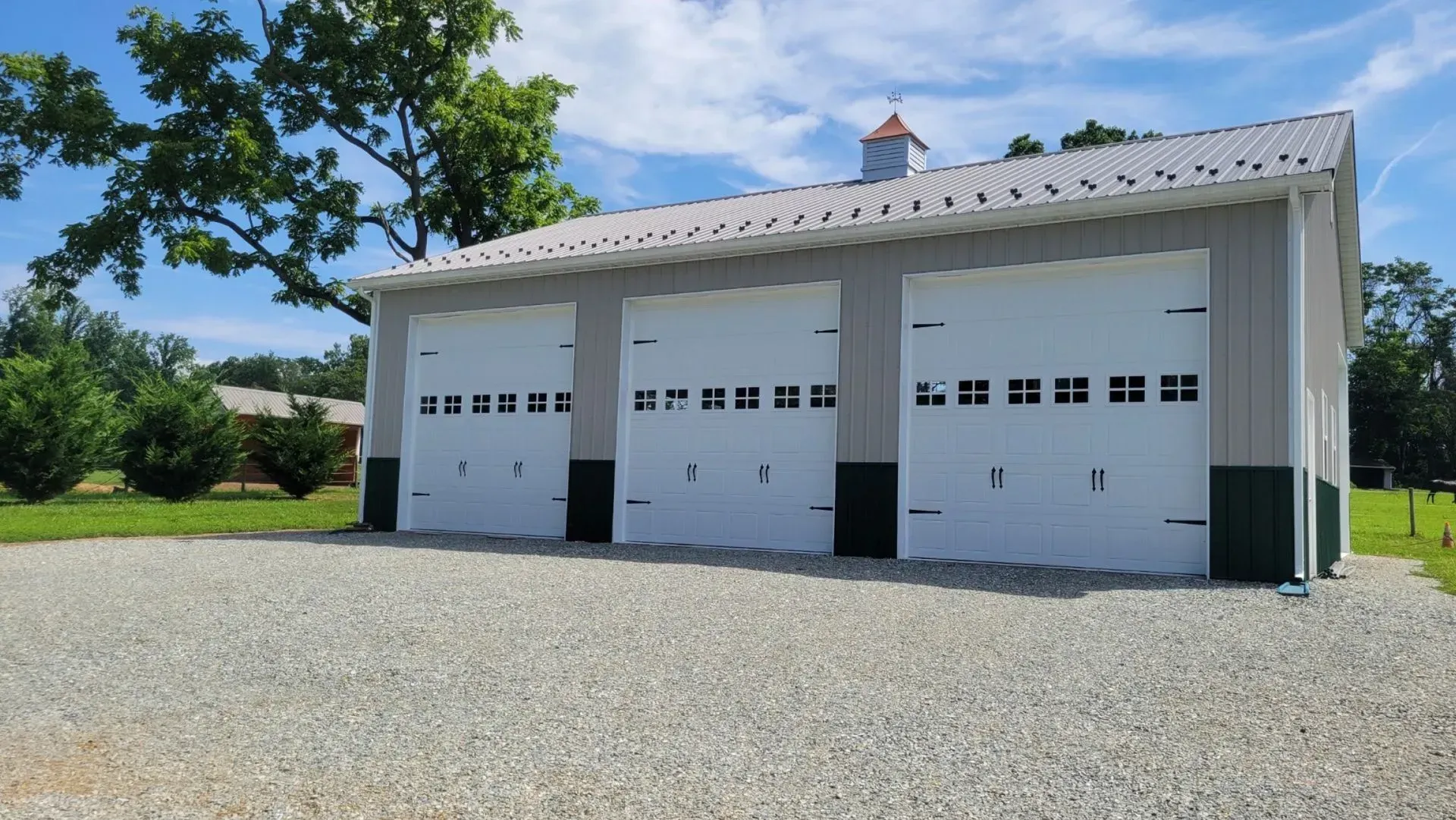 Three-bay garage with white doors and metal roof, gravel driveway, gray and green exterior, under a blue sky.