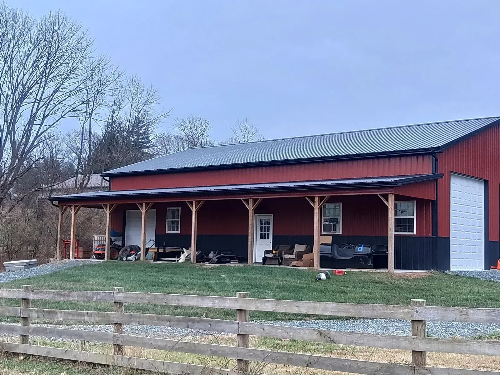 Red and black barn with a porch, metal roof, and garage door; set on a grassy hill with a wooden fence.
