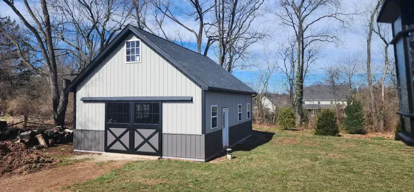 A gray barn with a dark roof and black doors sits on a grassy yard.