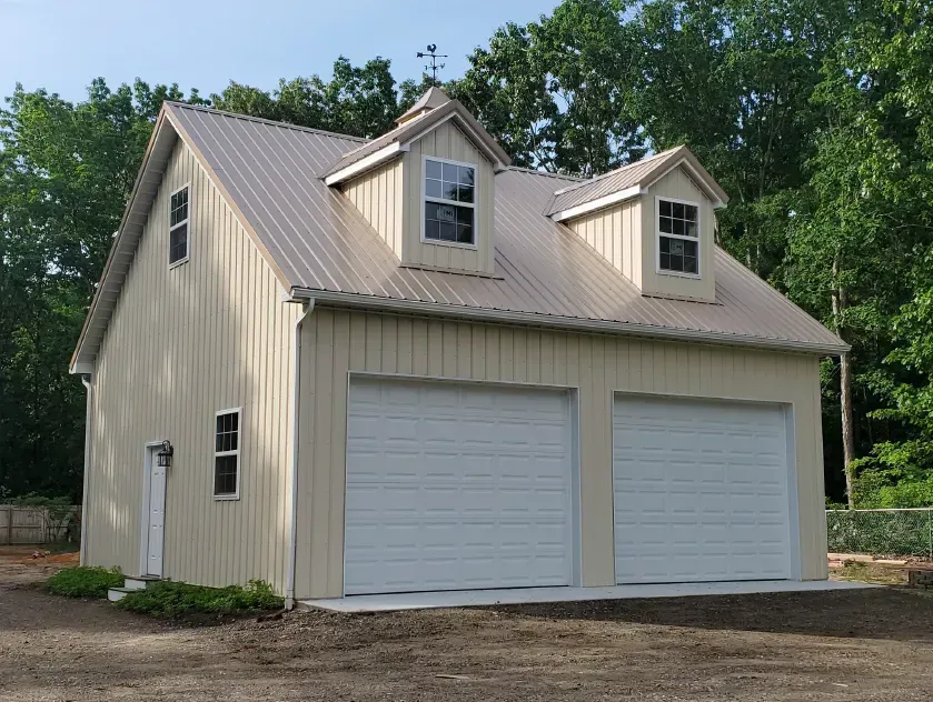 Two-story tan garage with a metal roof and two garage doors. Features dormer windows and a small side door.