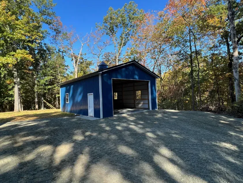 Blue barn with open garage door on gravel drive, surrounded by autumn trees.