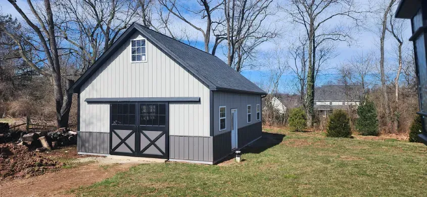 A light gray barn with black trim and doors, located on a grassy lawn with trees in the background.