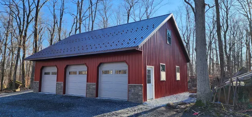 Red three-car garage with stone accents, a gray roof, and a gravel driveway, in a wooded area.