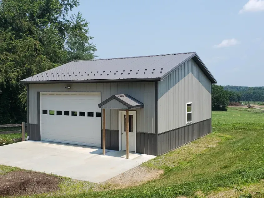 Gray and dark gray metal building with a garage door and small entry under a porch, sitting on a concrete pad.