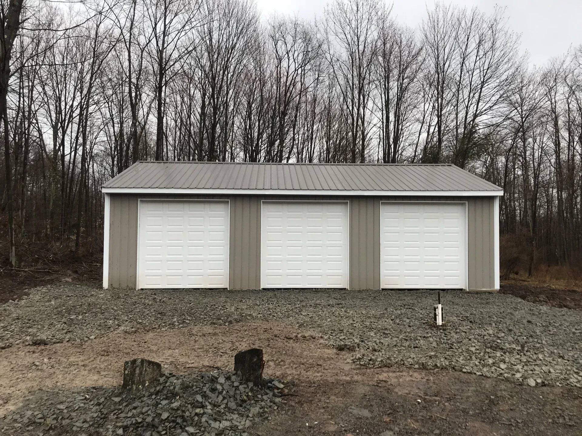Three-bay garage with white doors and gray siding, built on gravel, with a metal roof. Bare trees in background.