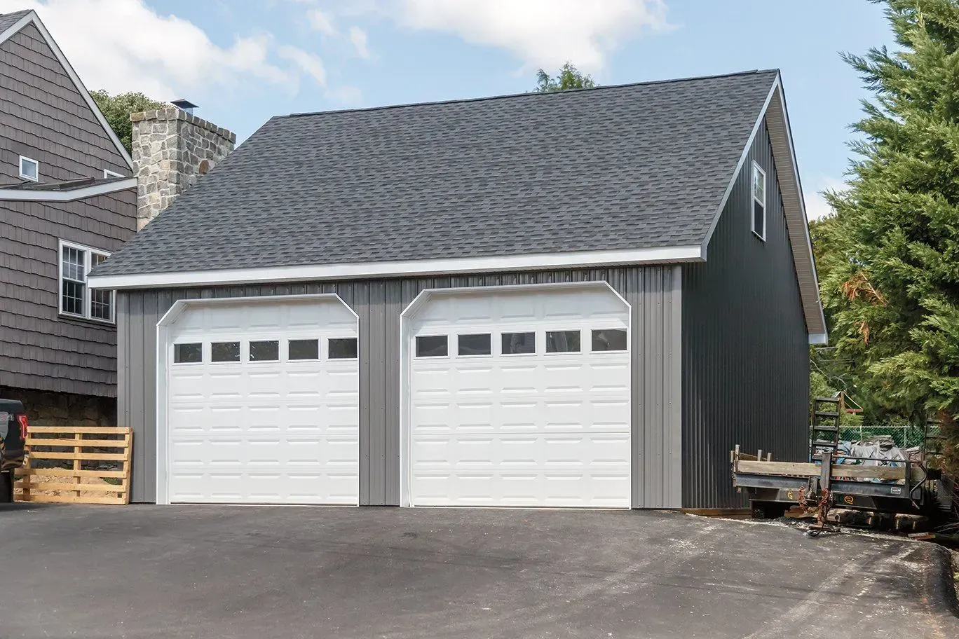 Two-car gray garage with white garage doors and a dark gray roof, next to a house and trees.