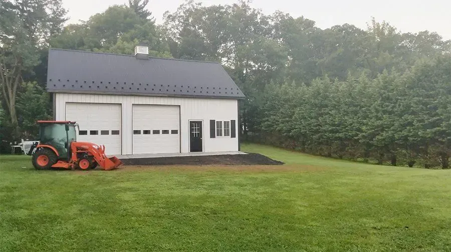 White building with black roof; two garage doors, tractor, and lush green lawn.