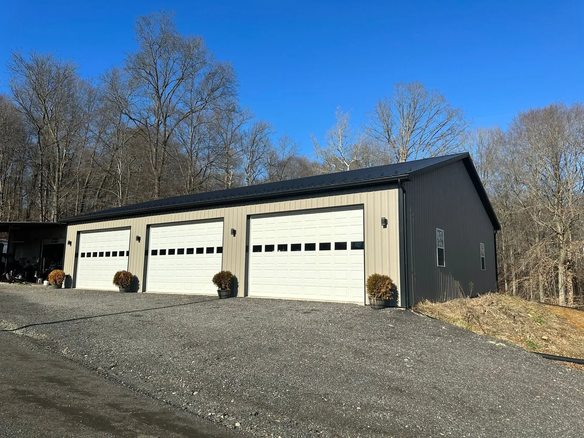 Three-bay garage with white doors, gray siding, and a black roof, set on a gravel driveway under a blue sky.