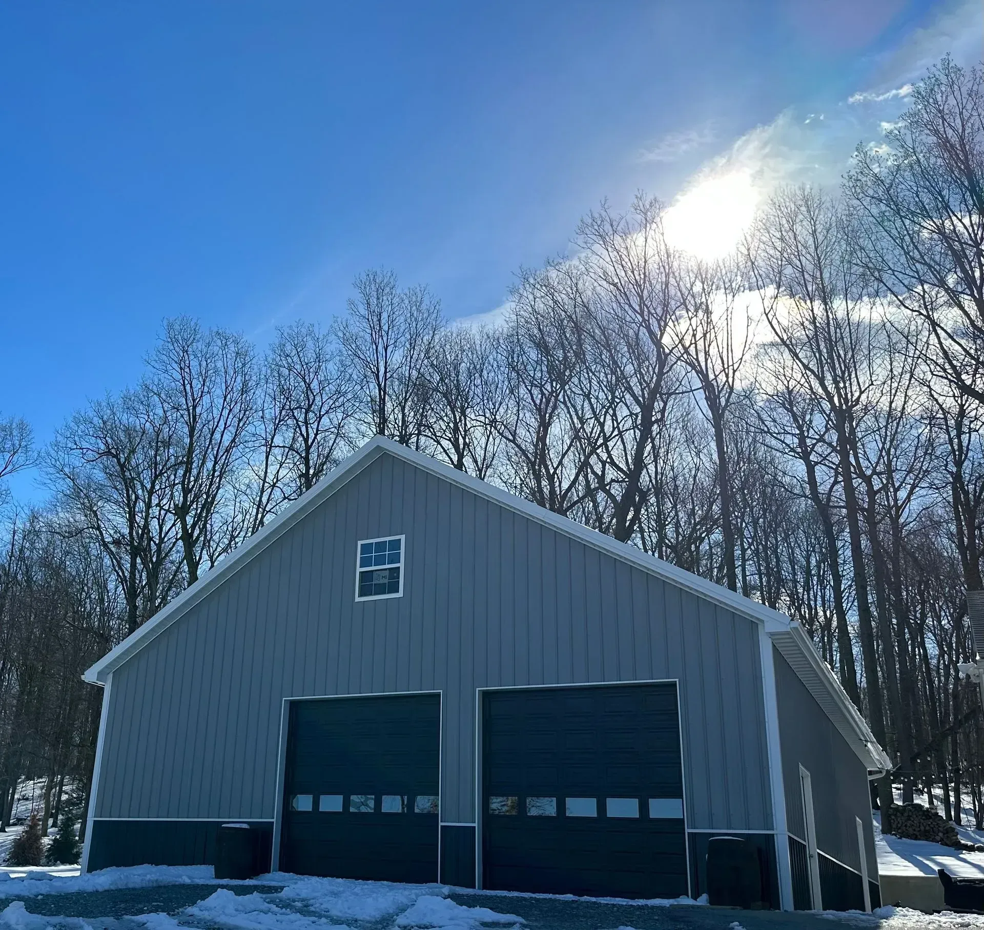 Gray metal barn with two garage doors, set against a background of bare trees and a bright blue sky.