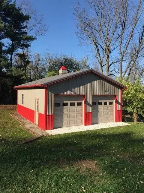 Metal garage with red trim, two garage doors, and tan siding, on a grassy lawn.