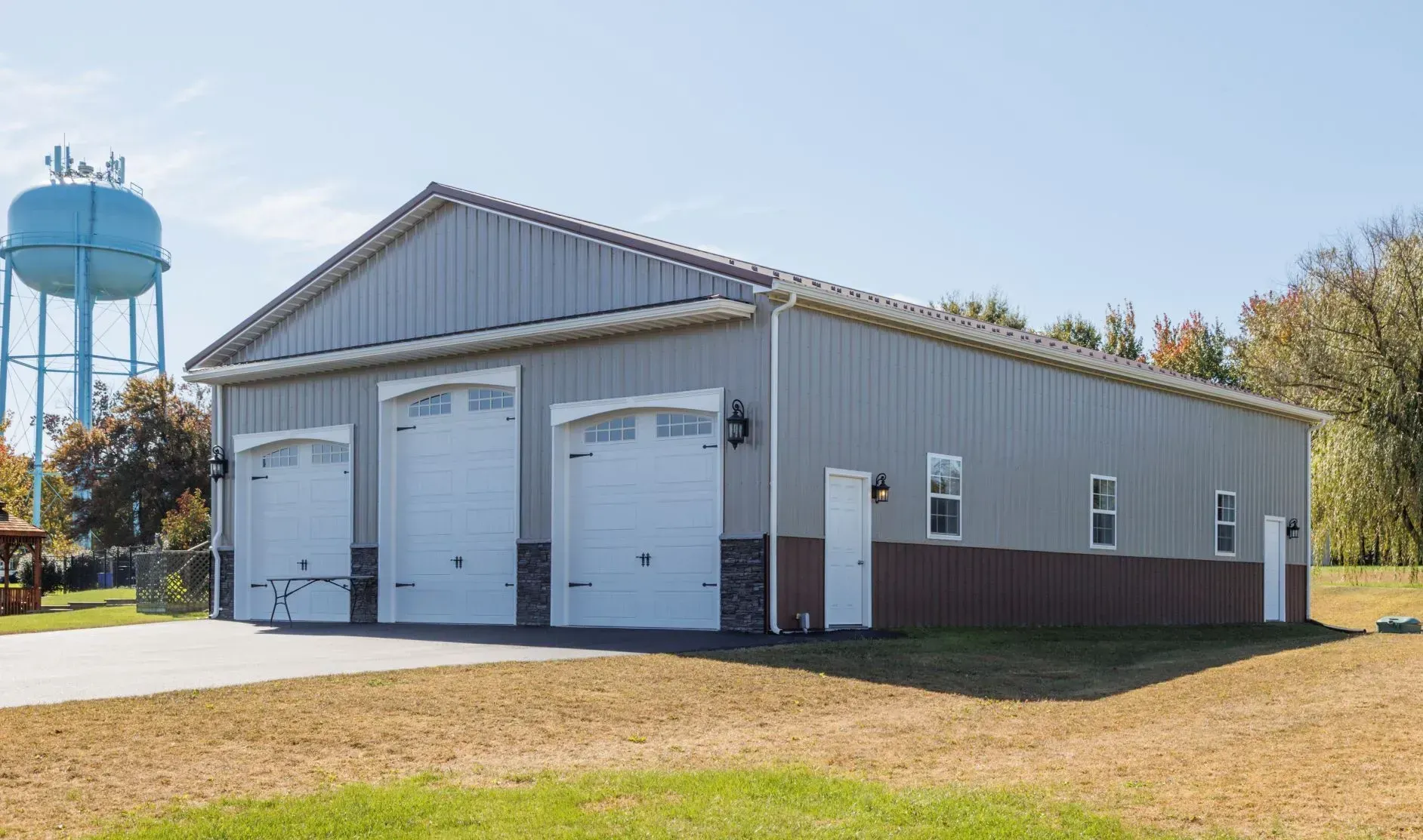 Three-bay metal garage with white doors, brown wainscoting, and a blue water tower in the background.