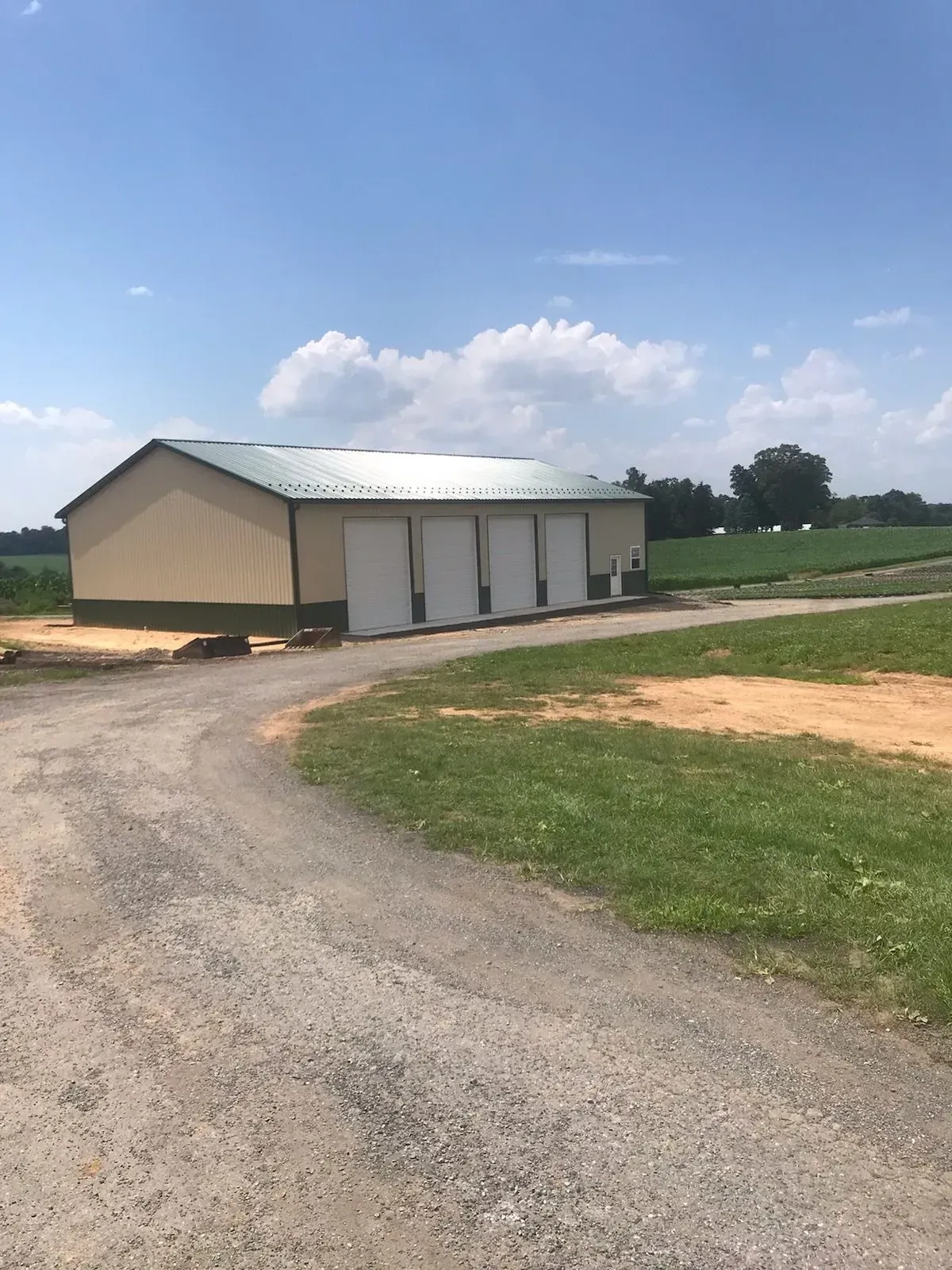 Tan and green metal building with white doors on a gravel road, fields in the background under a blue sky.