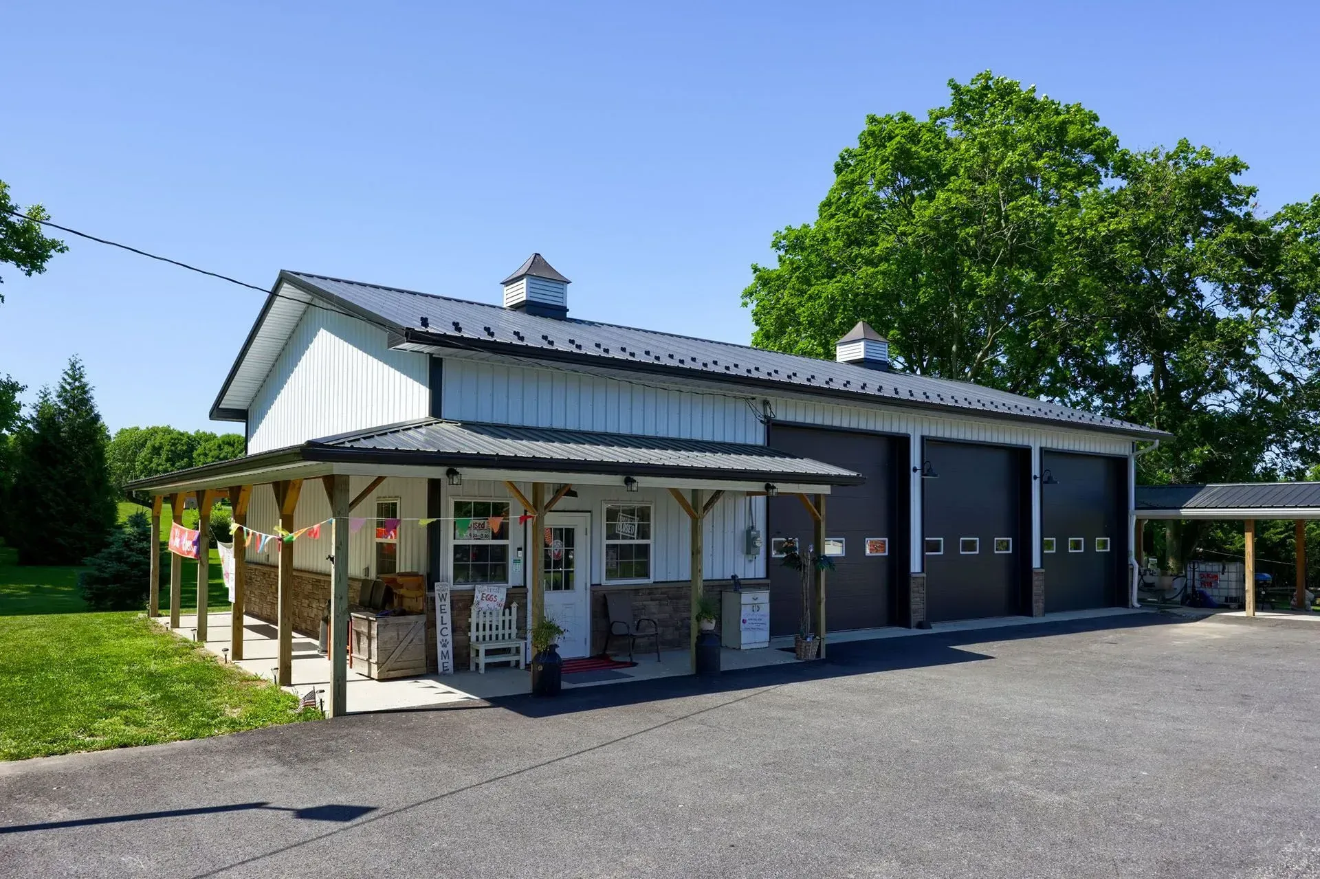 White and black barn-style building with a porch, garage doors, and a paved parking area on a sunny day.