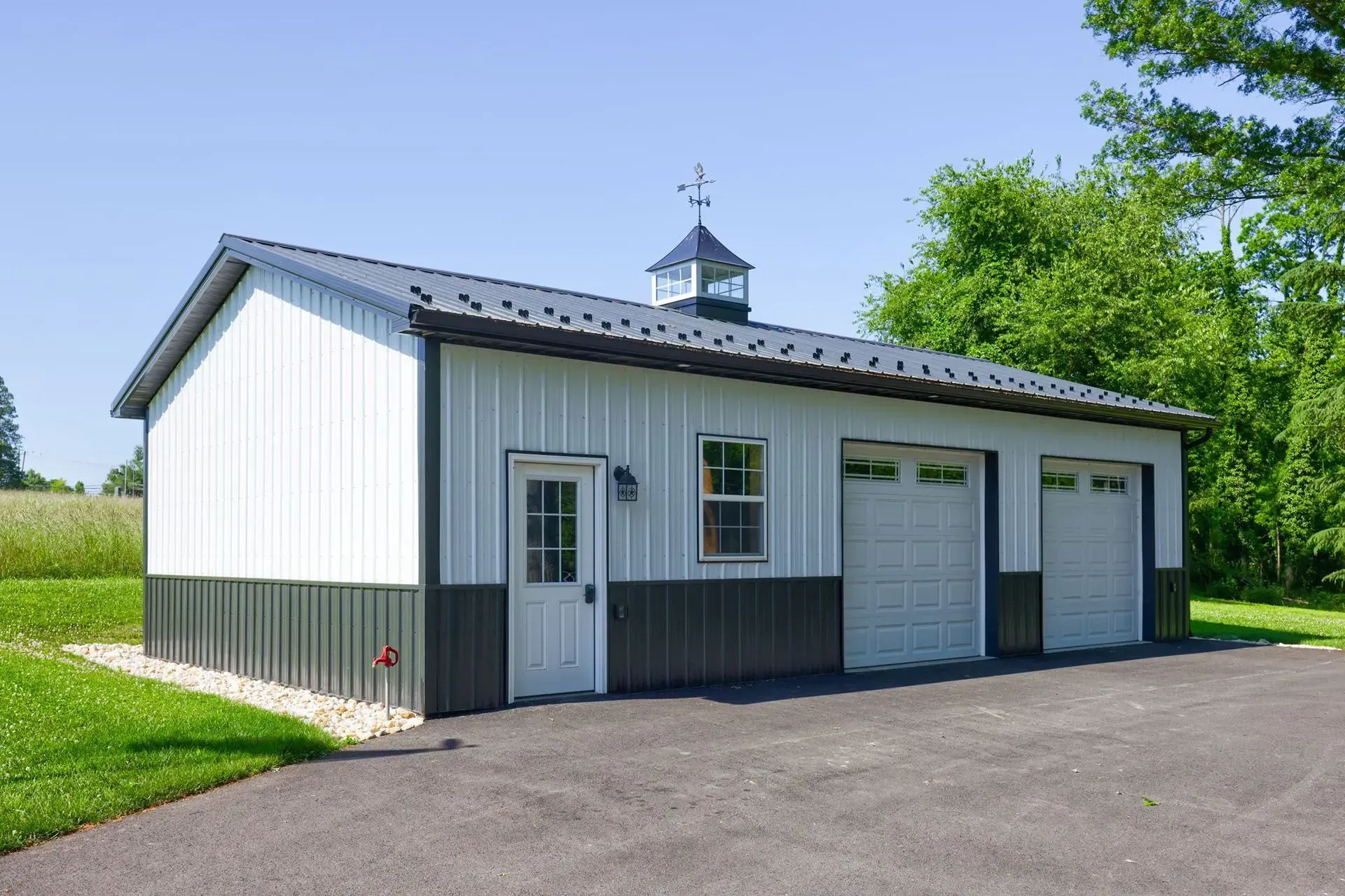 White and gray metal garage with three doors, a cupola, and a dark-colored lower portion; set on pavement with greenery.