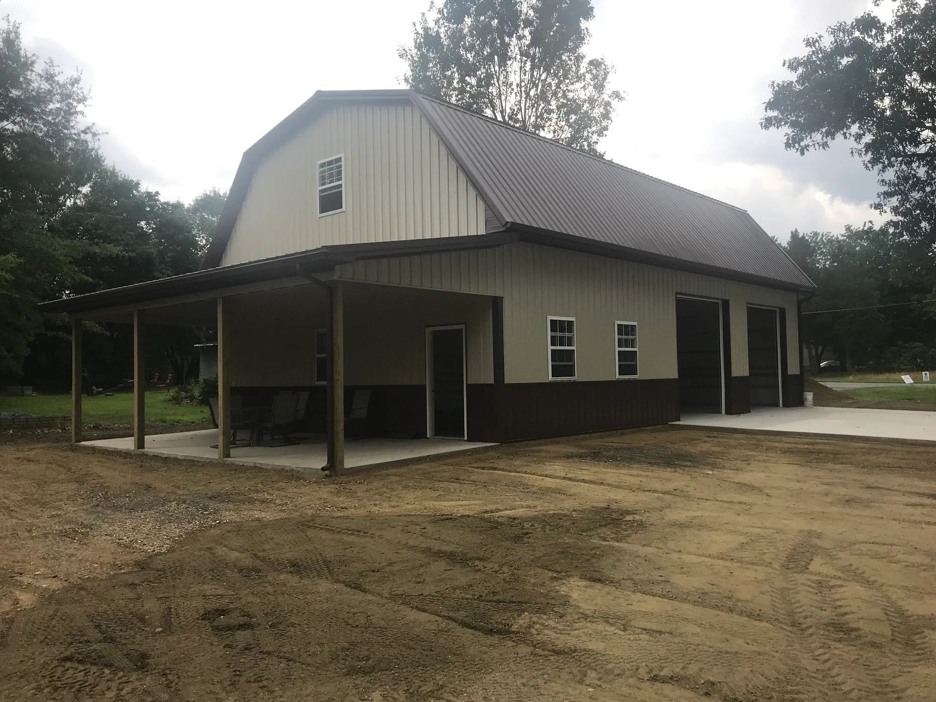 Tan and brown barn-style building with a covered porch and gravel driveway under an overcast sky.
