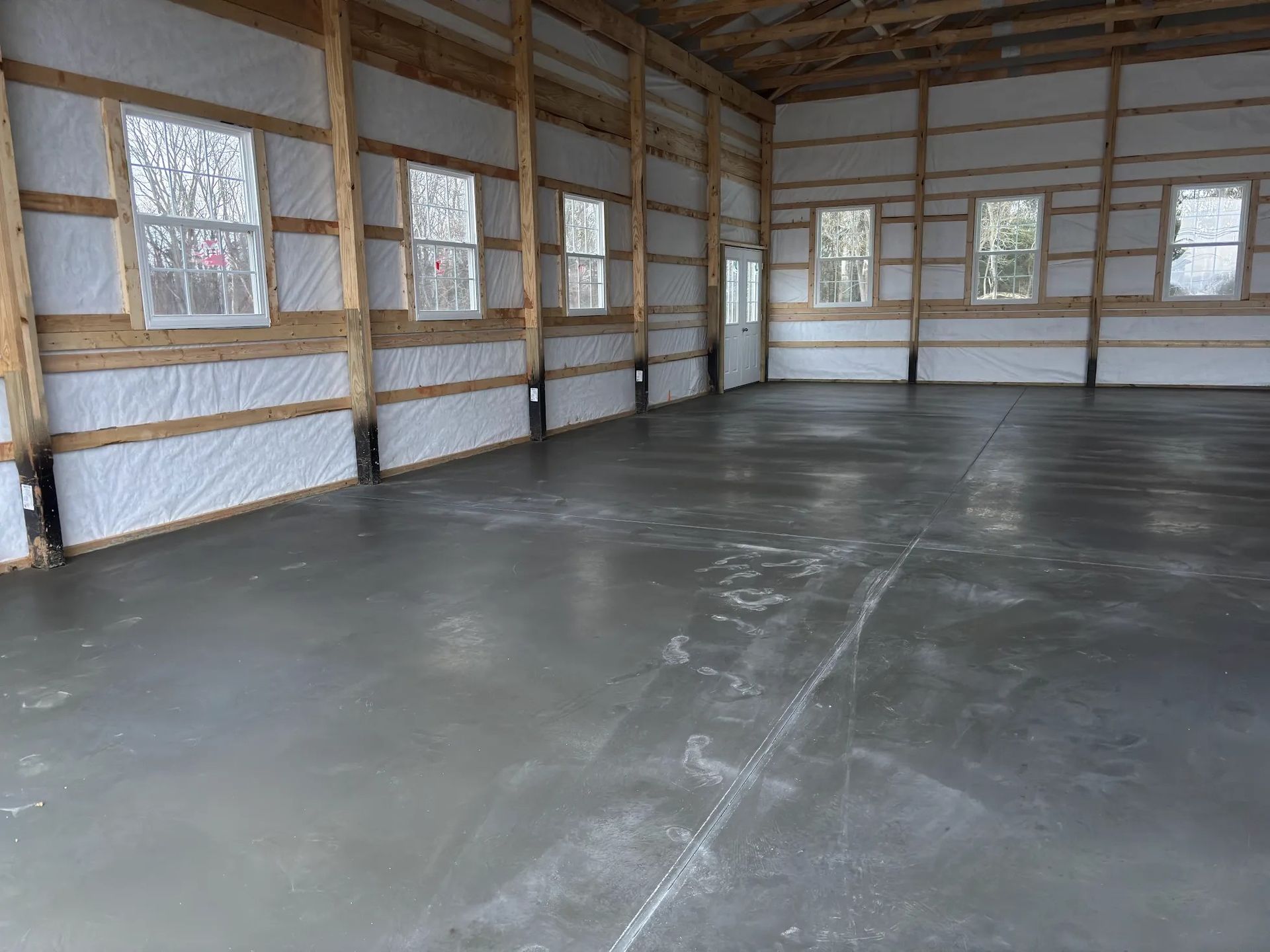 Interior of a large unfinished building with concrete floor and wooden framing; windows along the side walls.