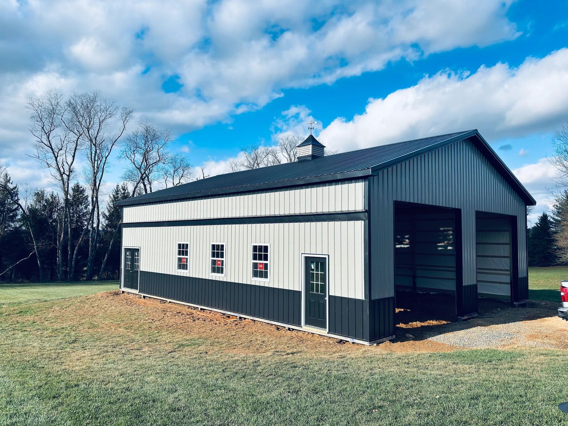 Steel-frame barn with gray and black panels. Two garage doors and multiple windows and a small cupola. Green grass and blue sky.