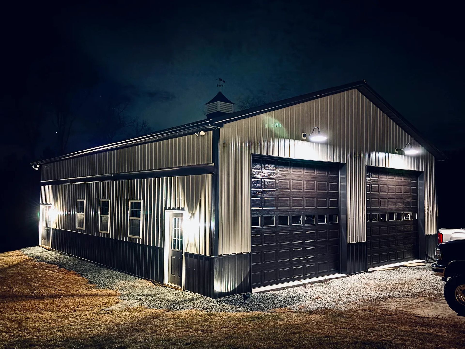 Metal-clad building with two garage doors lit up at night. Exterior lights and windows are visible.