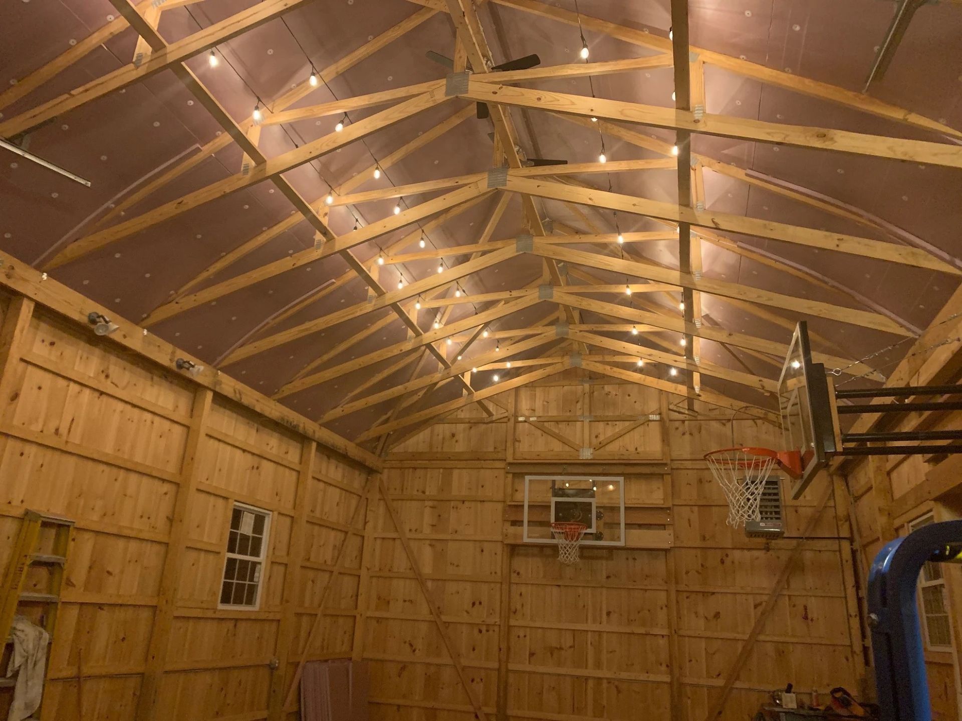 Wooden interior of a building, string lights hang from the rafters. Basketball hoops are mounted on the wall.