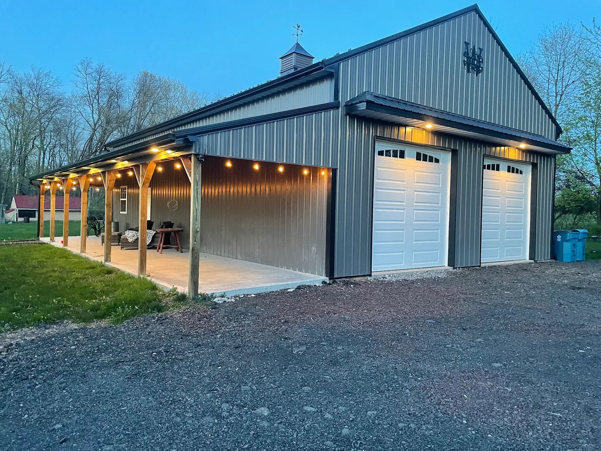 Gray metal barn with two garage doors and a covered porch lit by string lights.