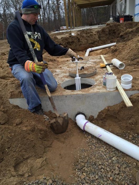 Worker in a trench installing white PVC drainage pipes around a concrete chamber.
