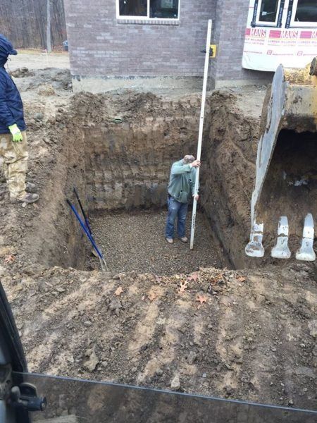 Deep excavation beside a brick house, with workers measuring the pit and an excavator bucket at right.