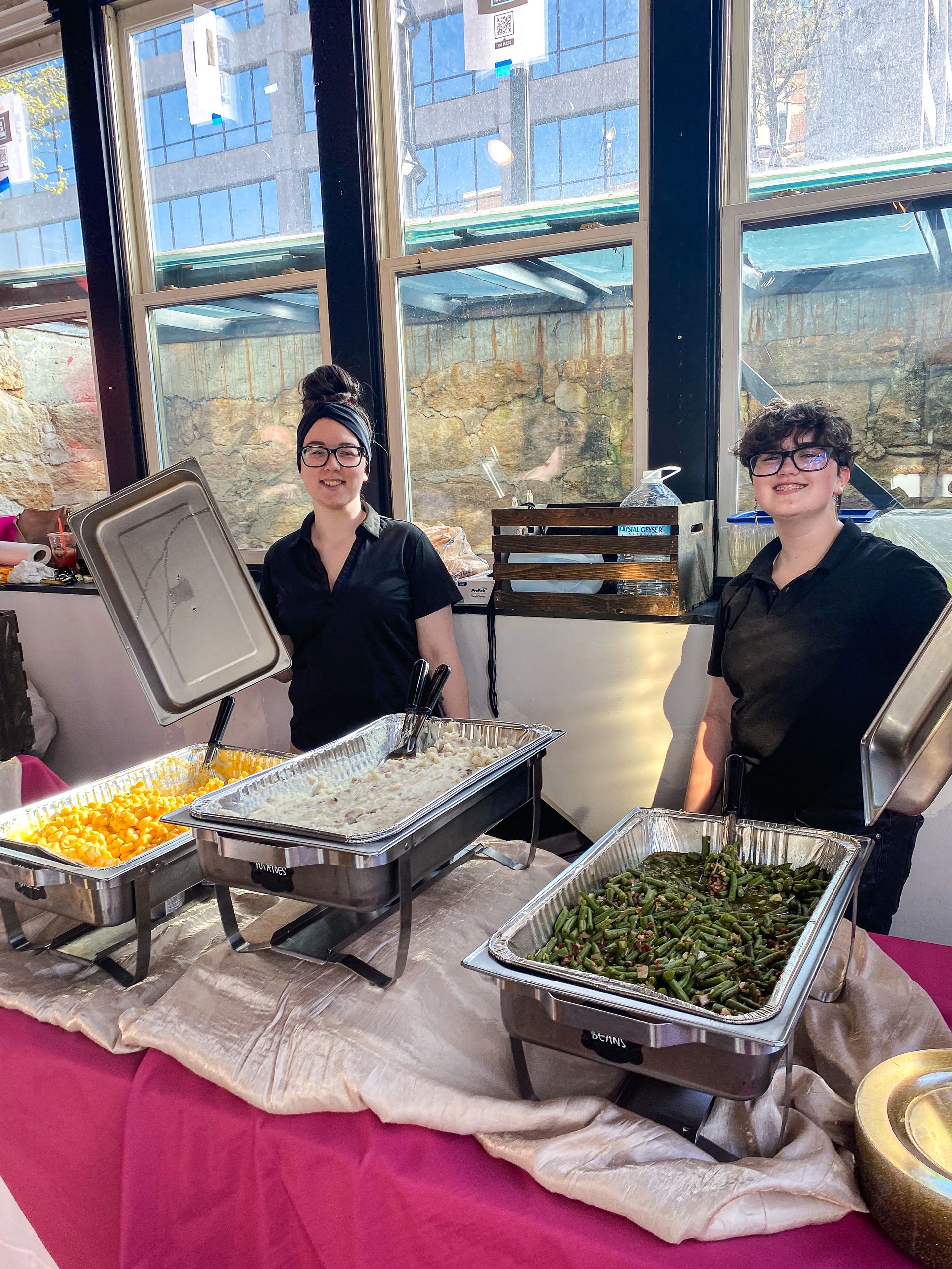 Two women are standing in front of a table filled with trays of food.