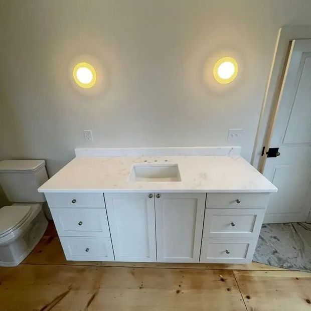 White bathroom vanity with marble countertop, cabinets, and three drawers. Two wall sconces and a toilet are visible.