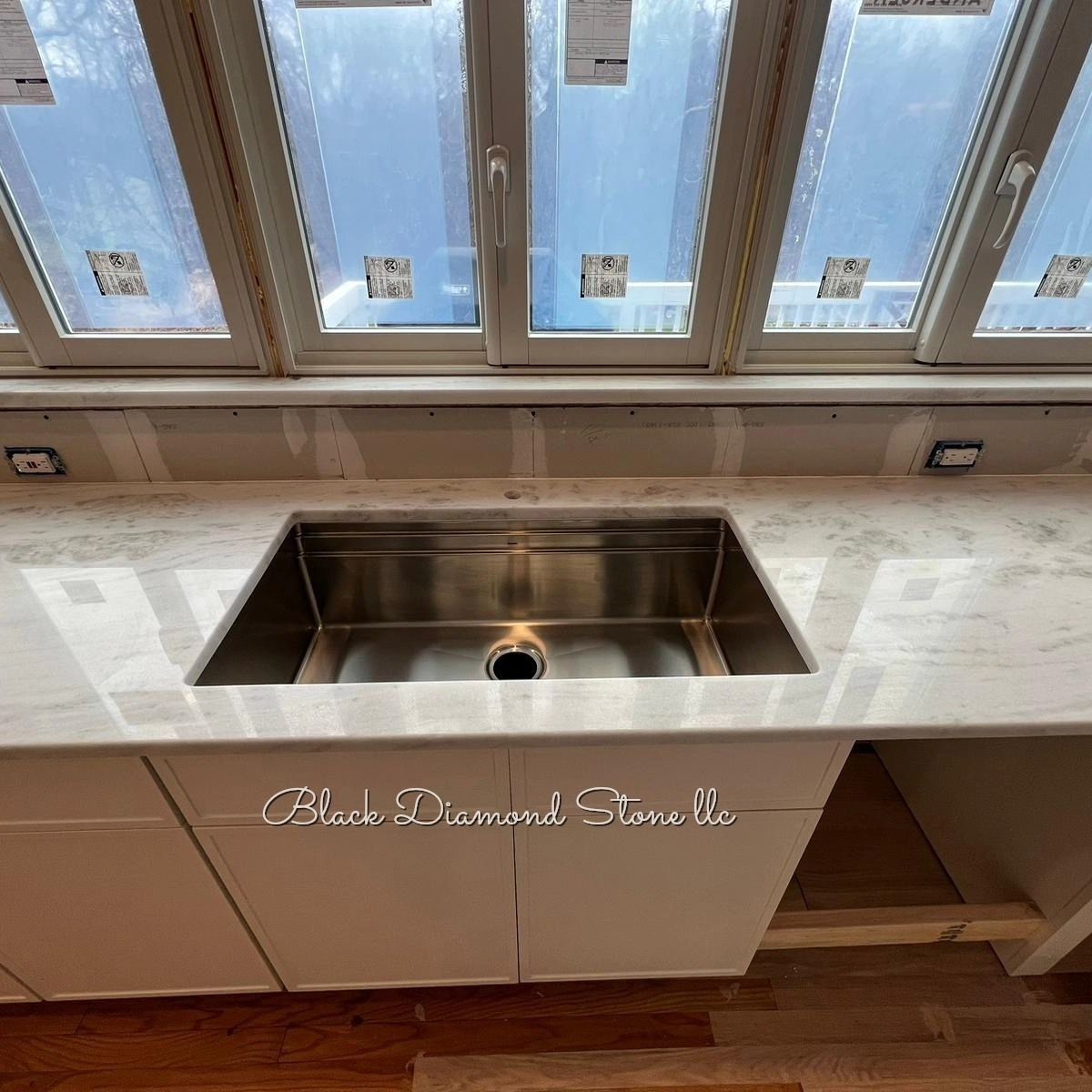 Stainless steel sink in white countertop under windows. Kitchen remodel in progress.
