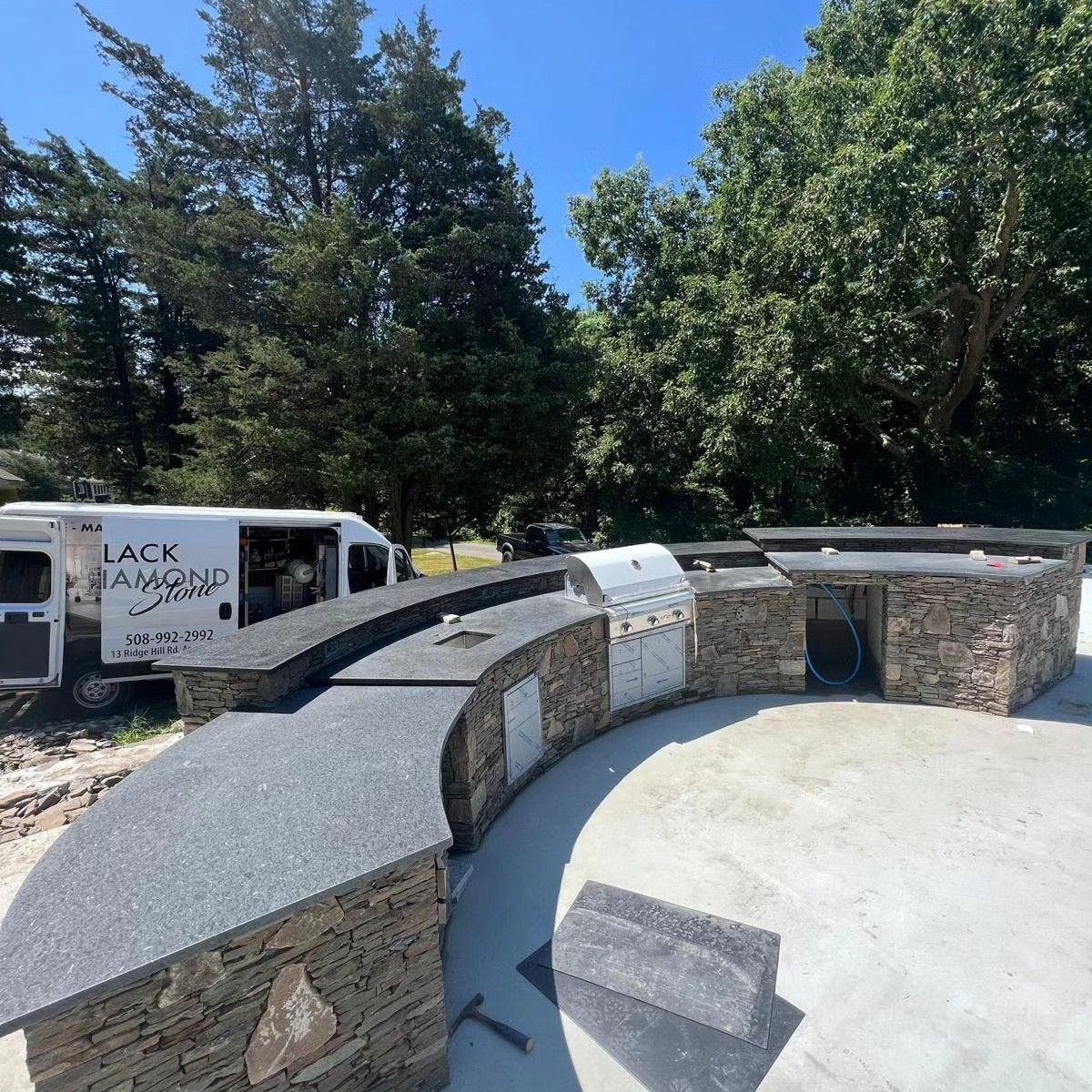 Outdoor kitchen with stone facing, granite countertops, and built-in grill, with van in the background.