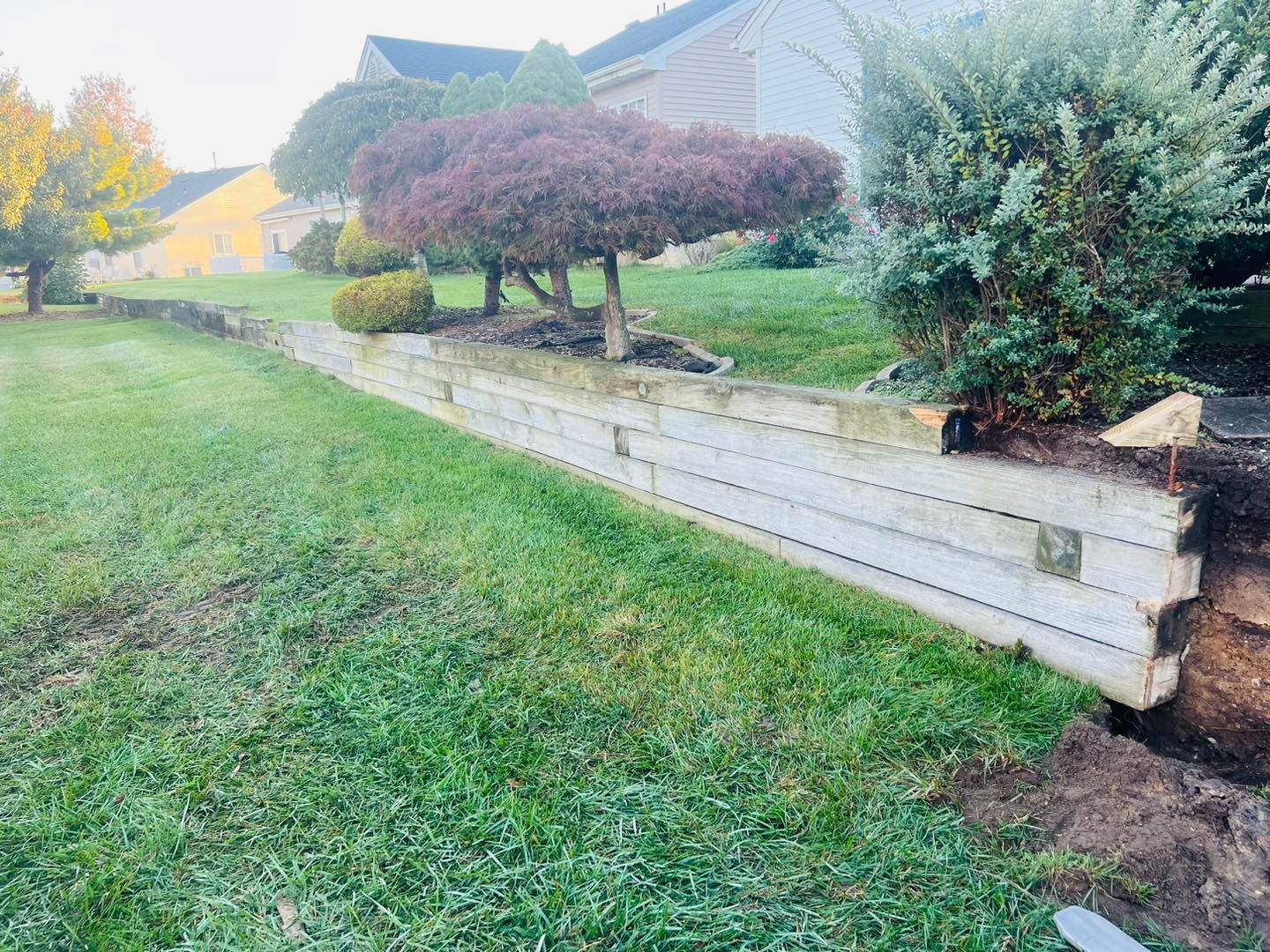 A wooden retaining wall is sitting in the middle of a lush green field next to a house.