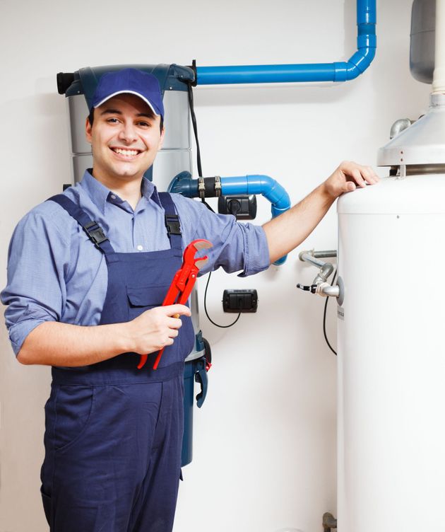 A smiling professional in a blue cap and work overalls holding a red wrench, standing next to a water heater.