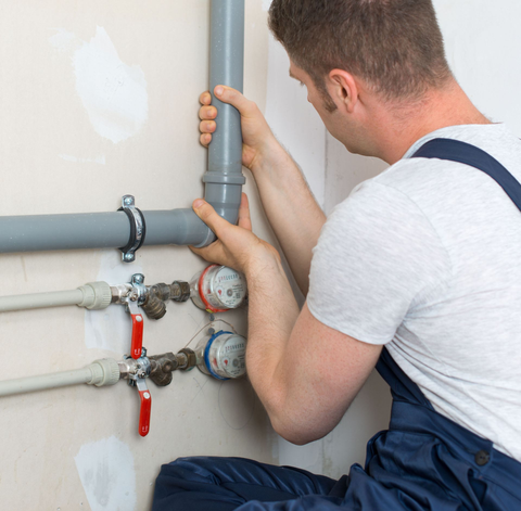 A plumber in blue overalls installs grey PVC pipes and water meters against a white wall.