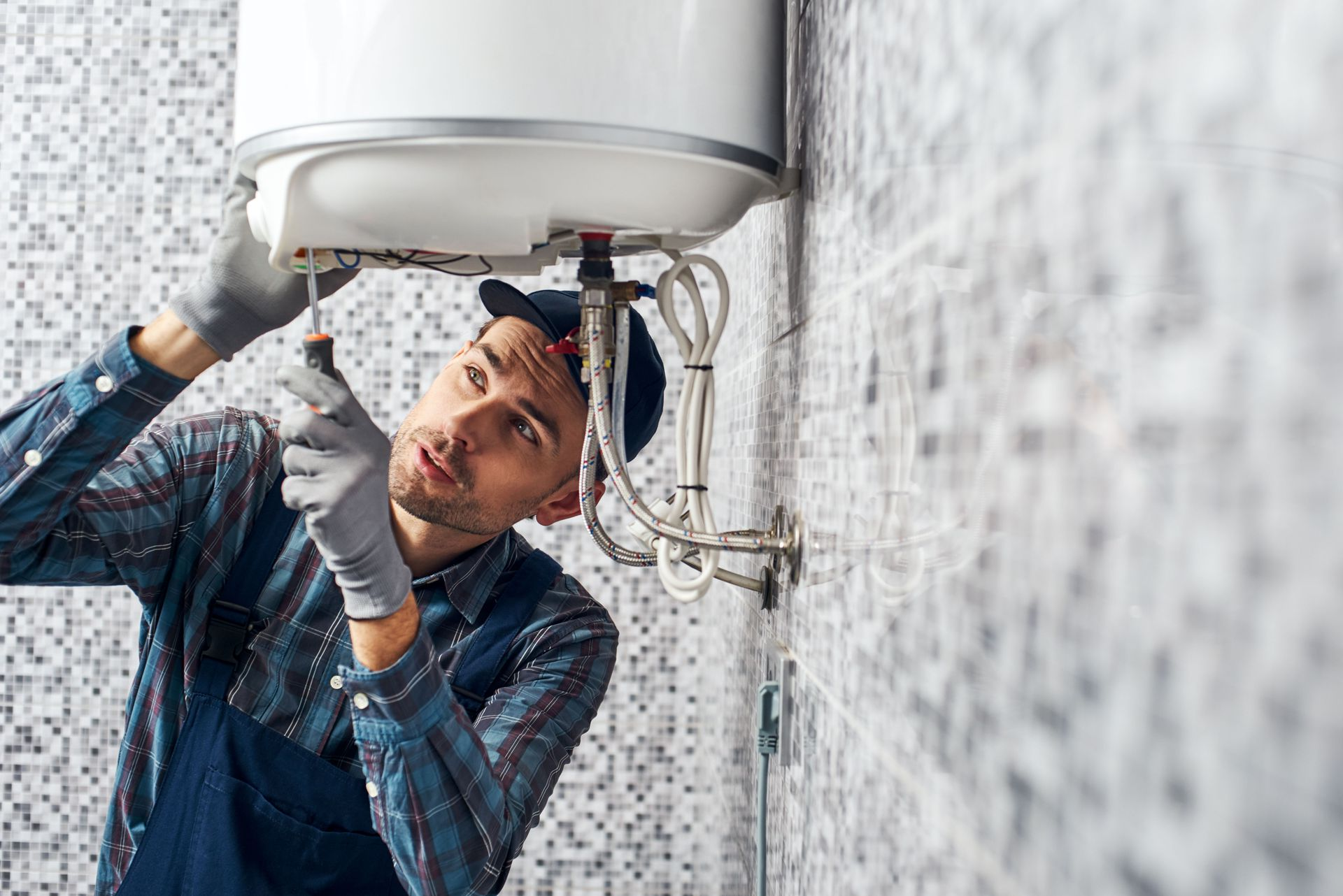 A worker in a plaid shirt and gloves repairs a white water heater mounted on a tiled wall.
