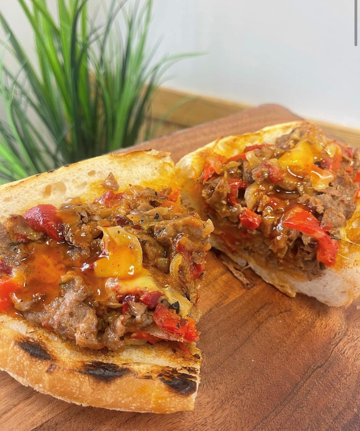 Close-up of a cheesesteak sandwich cut in half, revealing meat, peppers, and melted cheese on a toasted bun, on a wooden board.
