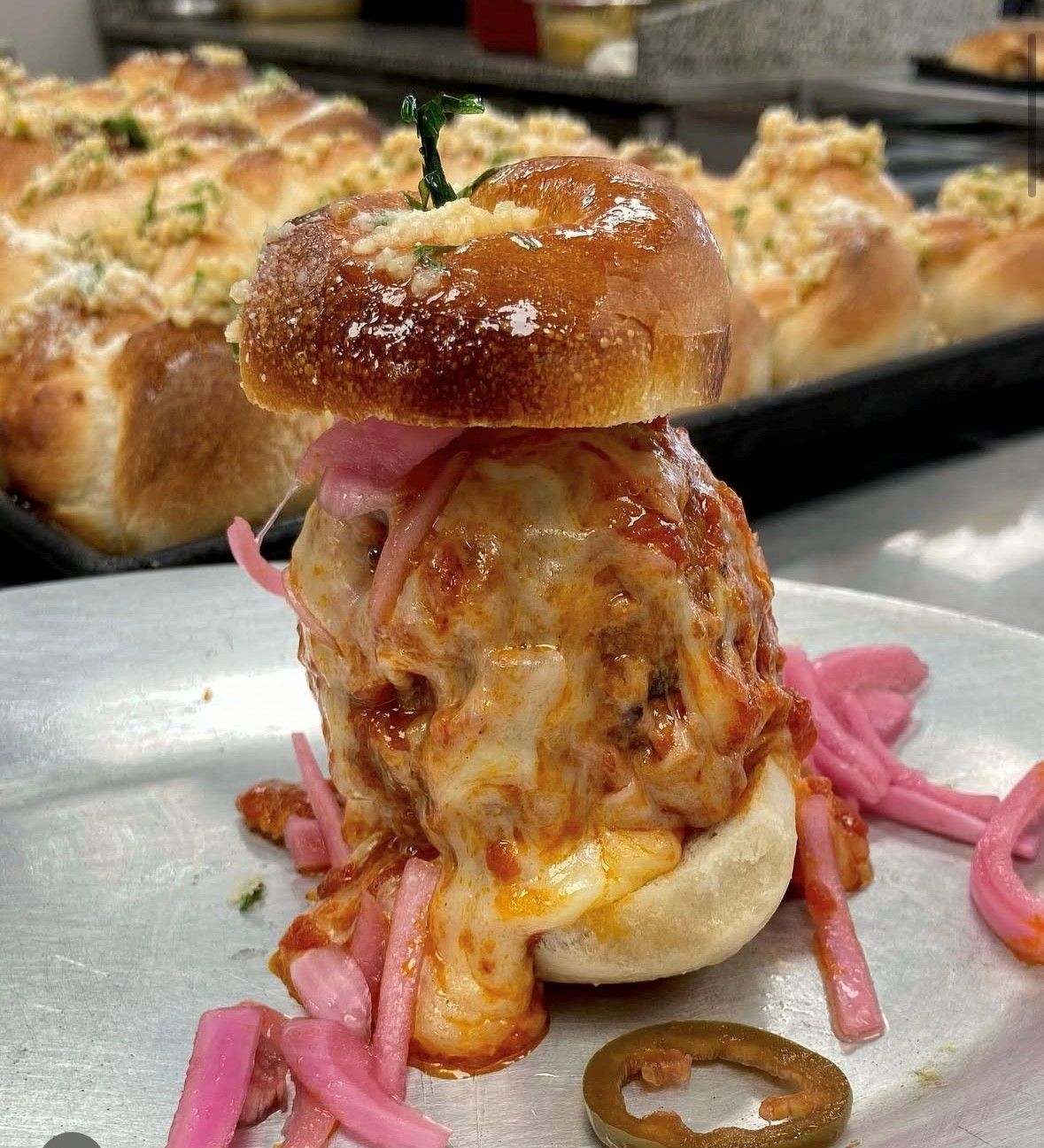 Meatball slider with cheese, sauce, pickled onions, and a brioche bun on a metal plate.