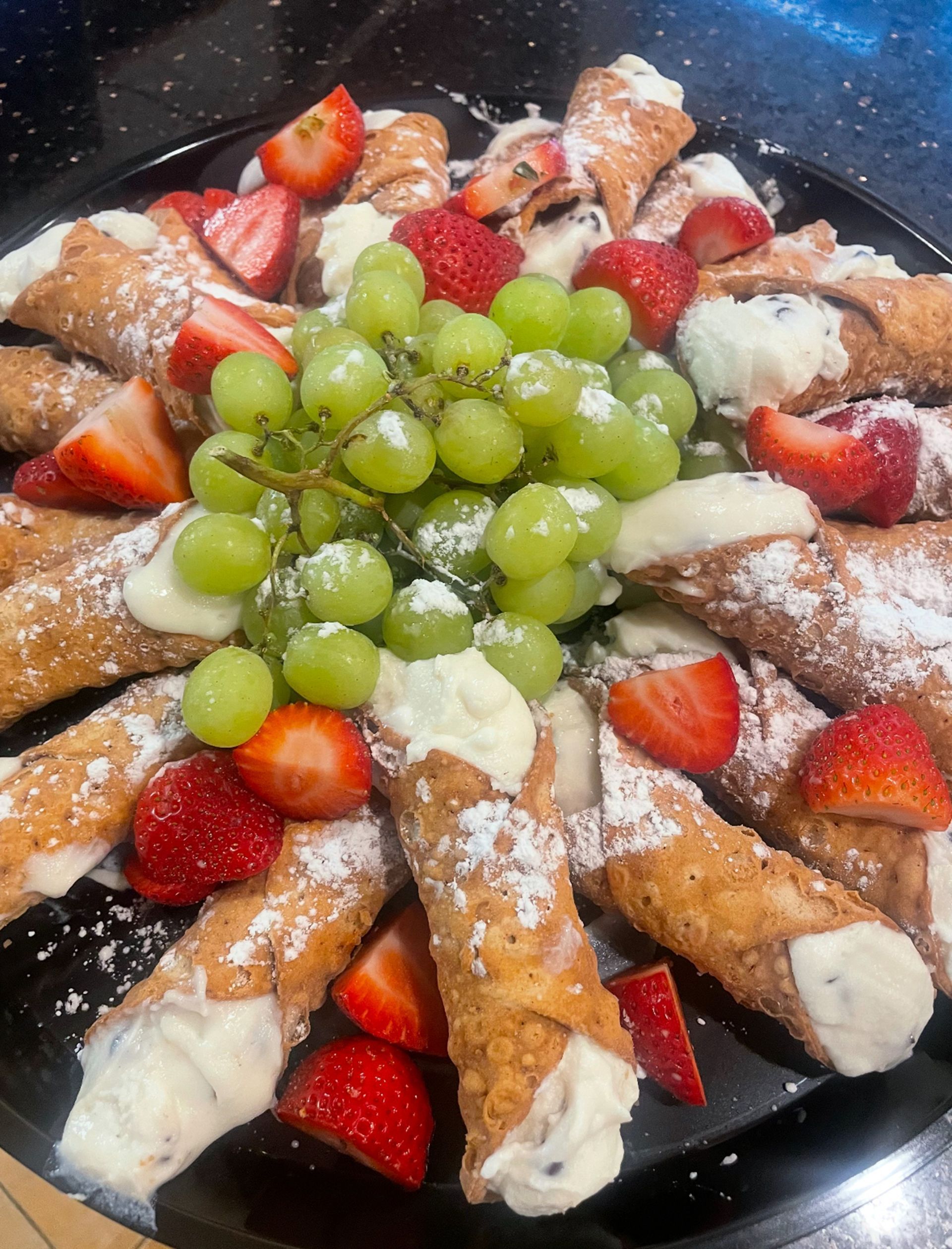 Cannoli pastries with creamy filling, garnished with strawberries and green grapes, dusted with powdered sugar.