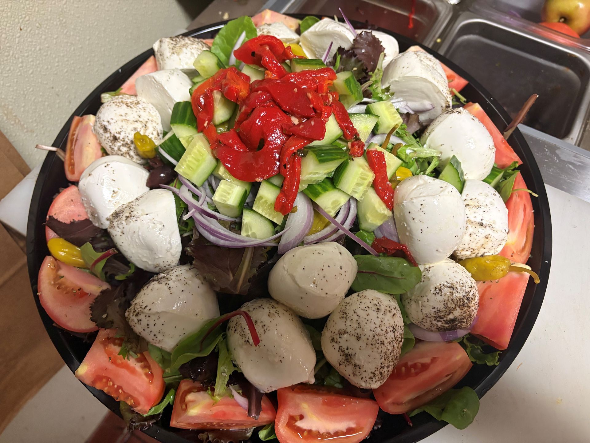Salad with mozzarella balls, tomatoes, cucumbers, peppers, and red onions in a black bowl.