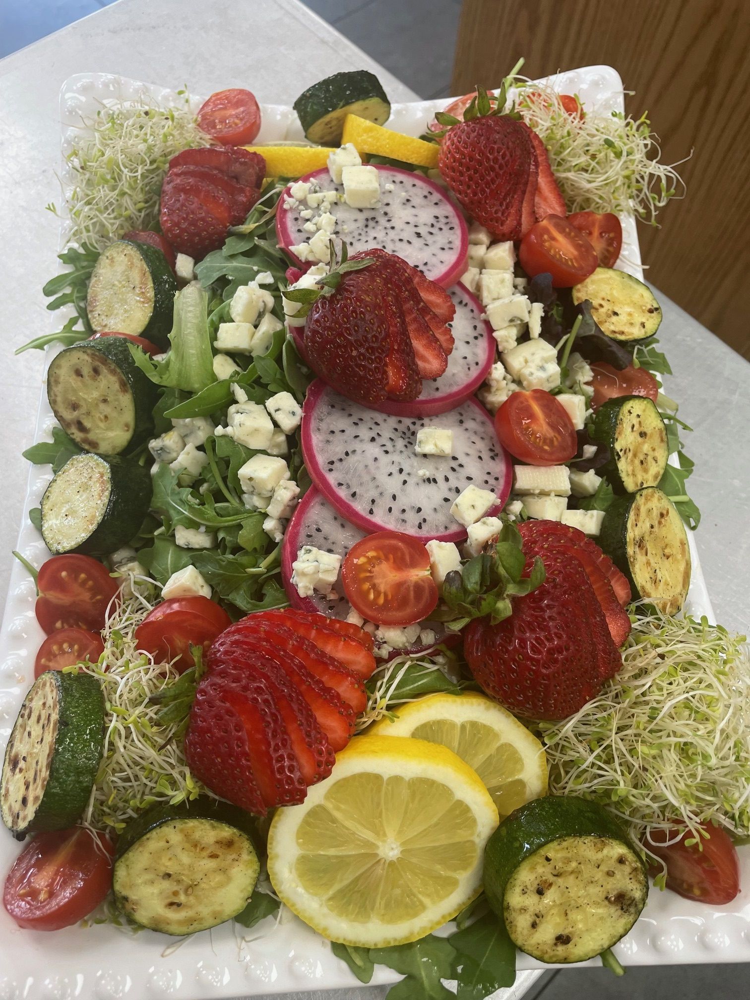 A salad with strawberries , dragon fruit , tomatoes , cucumbers , lemons and sprouts on a plate.