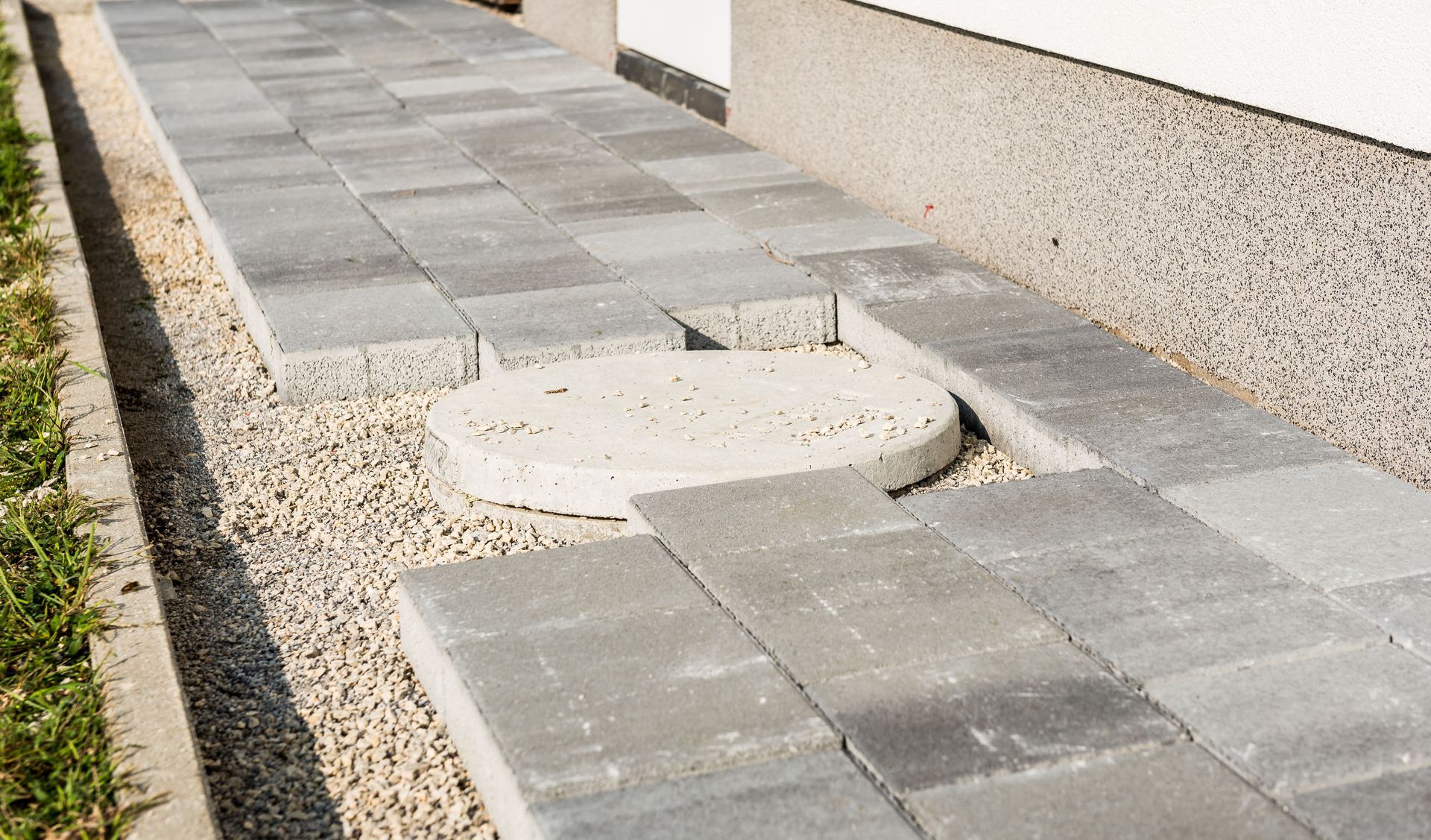 Paved sidewalk with a round utility access cover beside a gravel strip and building wall